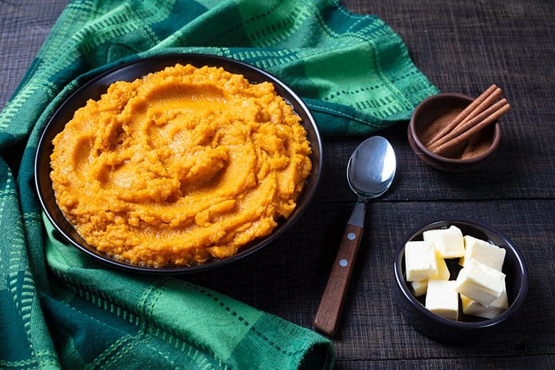 Mashed Sweet Potatoes in a medium serving plate, green tablecloth, spoon, cinnamon sticks and butter on its side