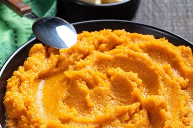 close up Mashed Sweet Potatoes in a medium serving plate, green tablecloth, spoon, and butter on its side