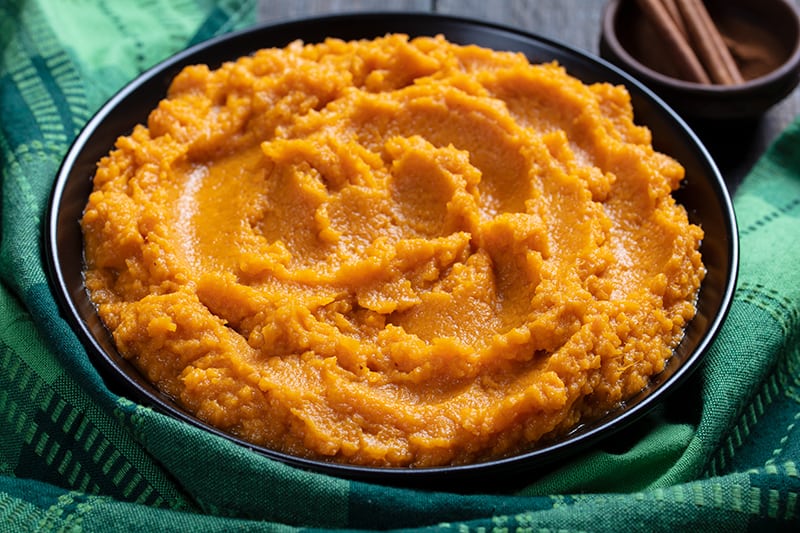 close up Mashed Sweet Potatoes in a medium serving plate, green tablecloth and cinnamon sticks on its background