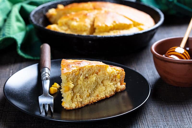 a slice of Sweet Honey Skillet Cornbread on black dessert plate with fork, whole skillet cornbread and honey on background