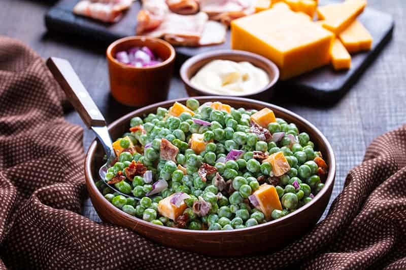 brown polka dot tablecloth underneath a bowl of Cheddar Bacon Green Pea Salad with a spoon on it, ingredients on its background