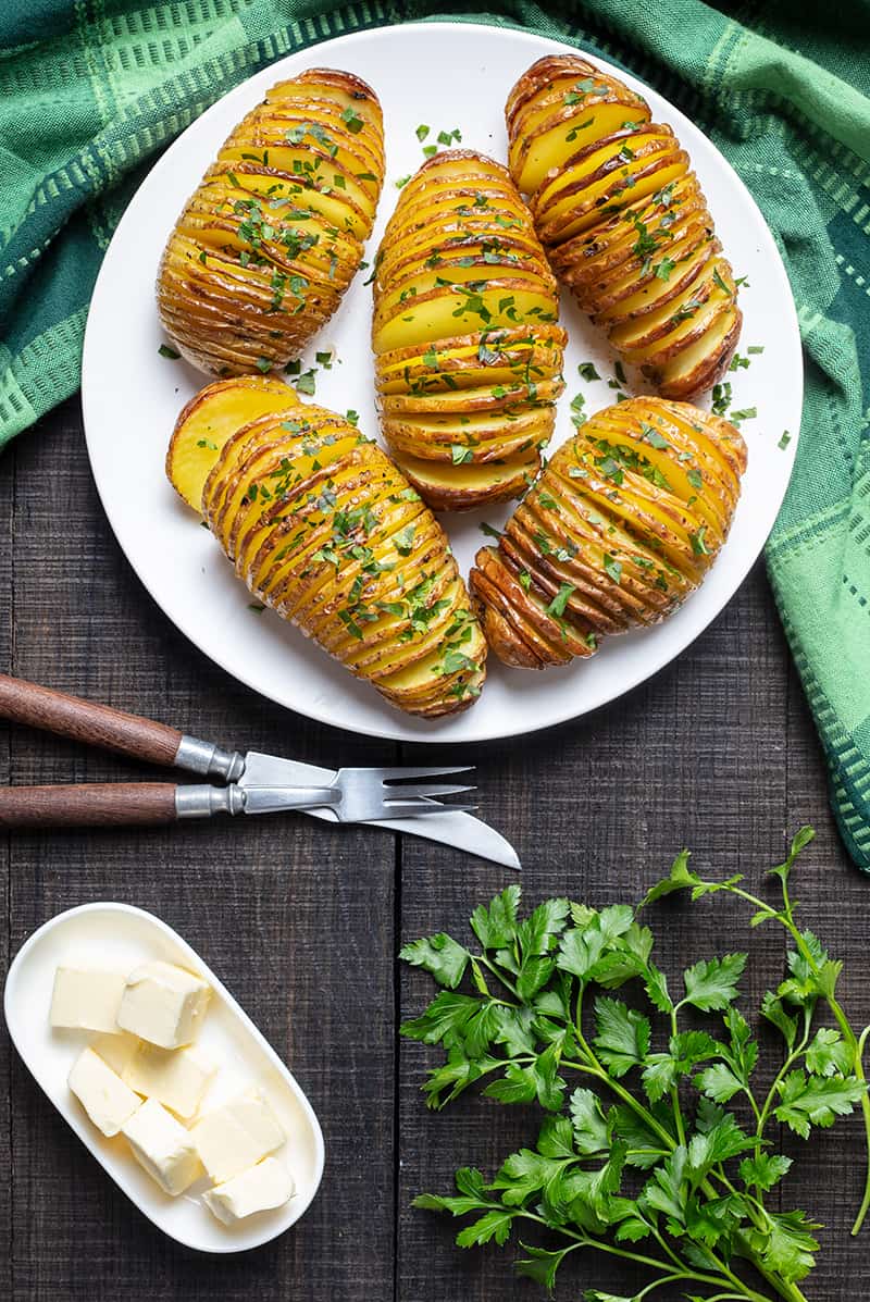 Final shot of roasted Hasselback Potatoes in white plate, green tablecloth parsley leaves butter bread knife and fork on side