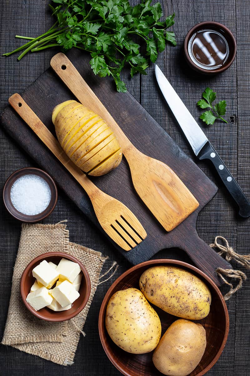 Hasselback Potato Ingredient Spread, cut potato in between wooden spoon on a wooden chopping board