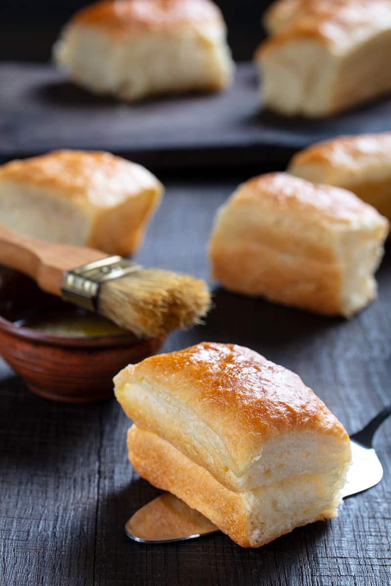 close up piece of soft buttery delicious Parker House Rolls, more Parker House Rolls and a basting brush on top of small bowl on its background