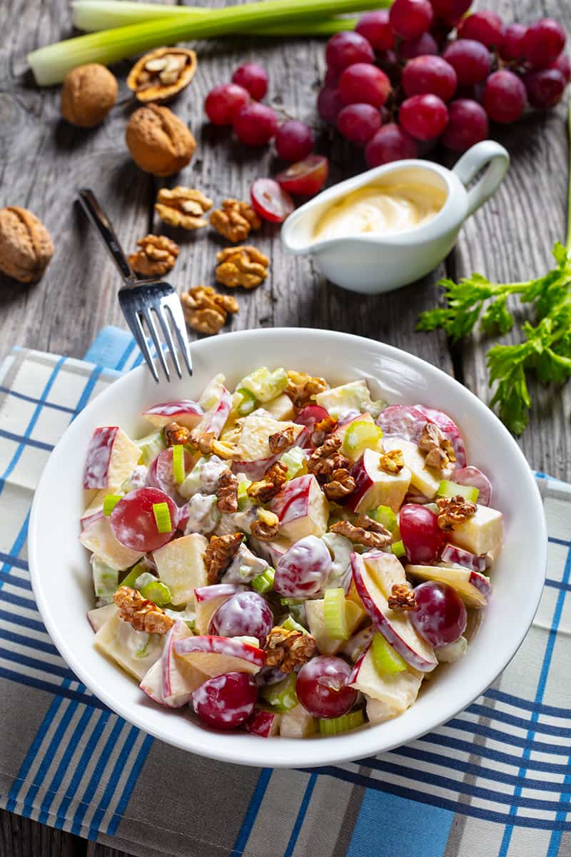 checkered blue tablecloth underneath white dessert bowl with Classic Waldorf Salad and fork on its side, ingredients on its background