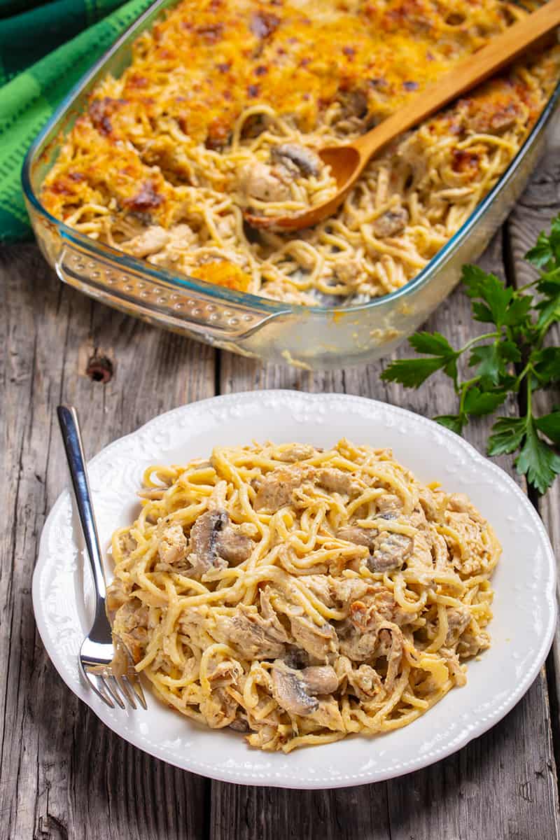 green tablecloth beside the glass baking dish of chicken tetrazzini with wooden spoon, some parsley leaves beside it and a white plate with some Chicken Tetrazzini and fork