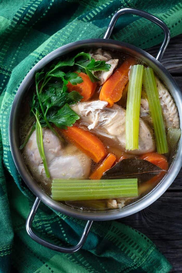 a green table cloth and boiling pan with Chicken broth made with vegetables and chicken breast , white soup bowl in a plate filled with chicken broth, salt and pepper shakers beside it