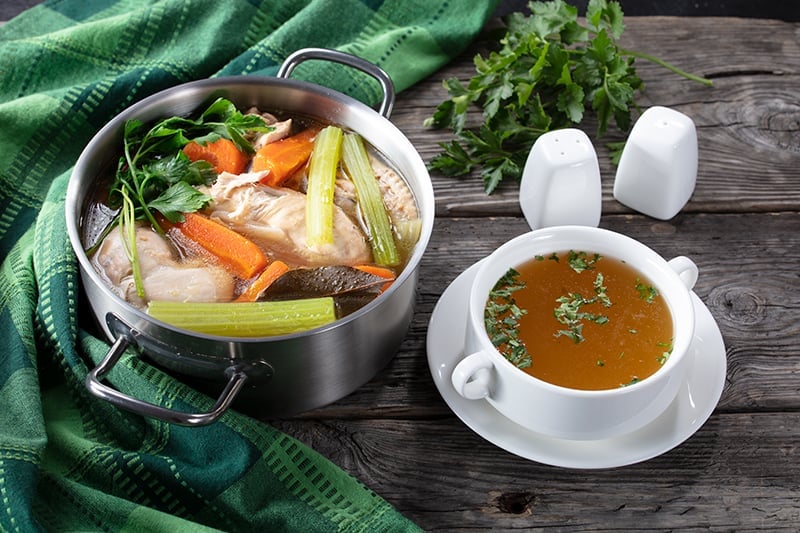 a green table cloth and boiling pan with Chicken broth made with vegetables and chicken breast , white soup bowl in a plate filled with chicken broth, salt and pepper shakers beside it