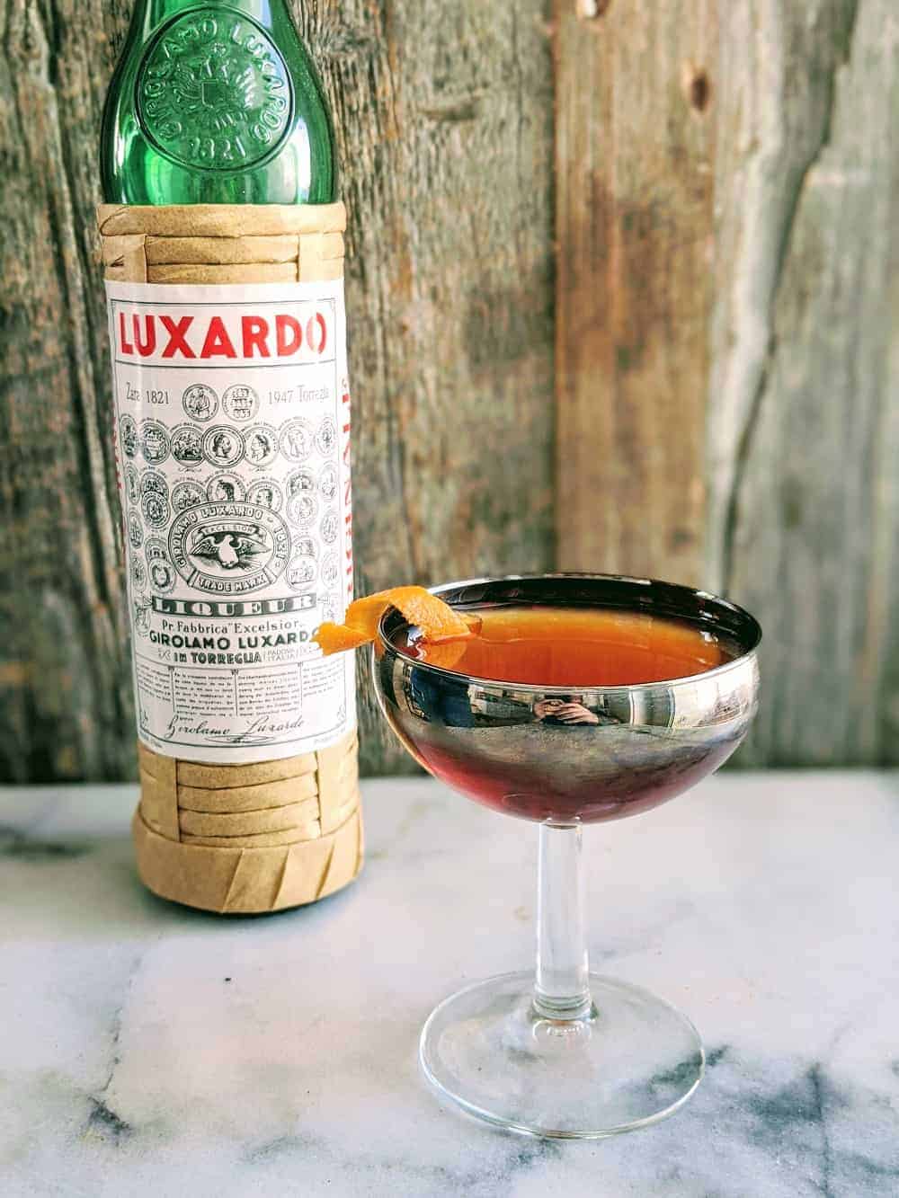 Martinez Cocktail in a silver coupe glass with a bottle of Luxardo behind it