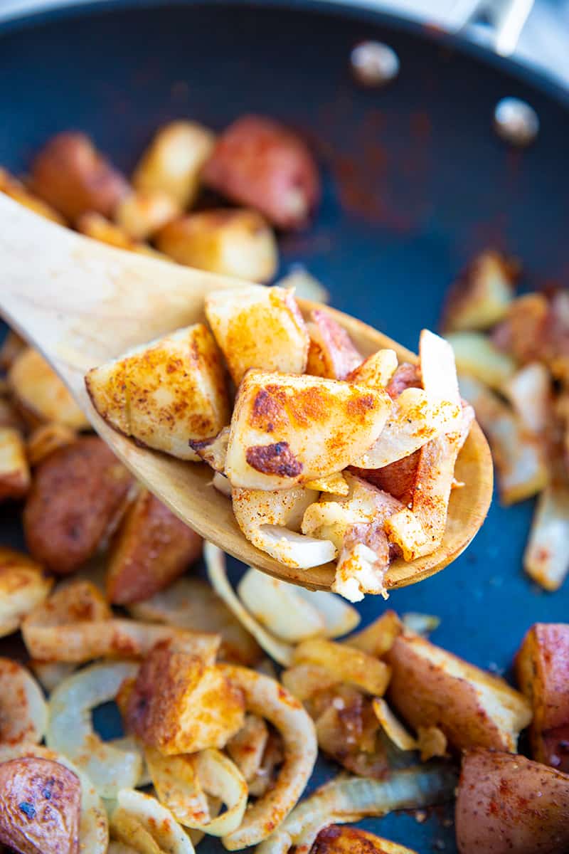 close up crispy pan fried potatoes and onions with paprika in a medium sized skillet with wooden spoon 