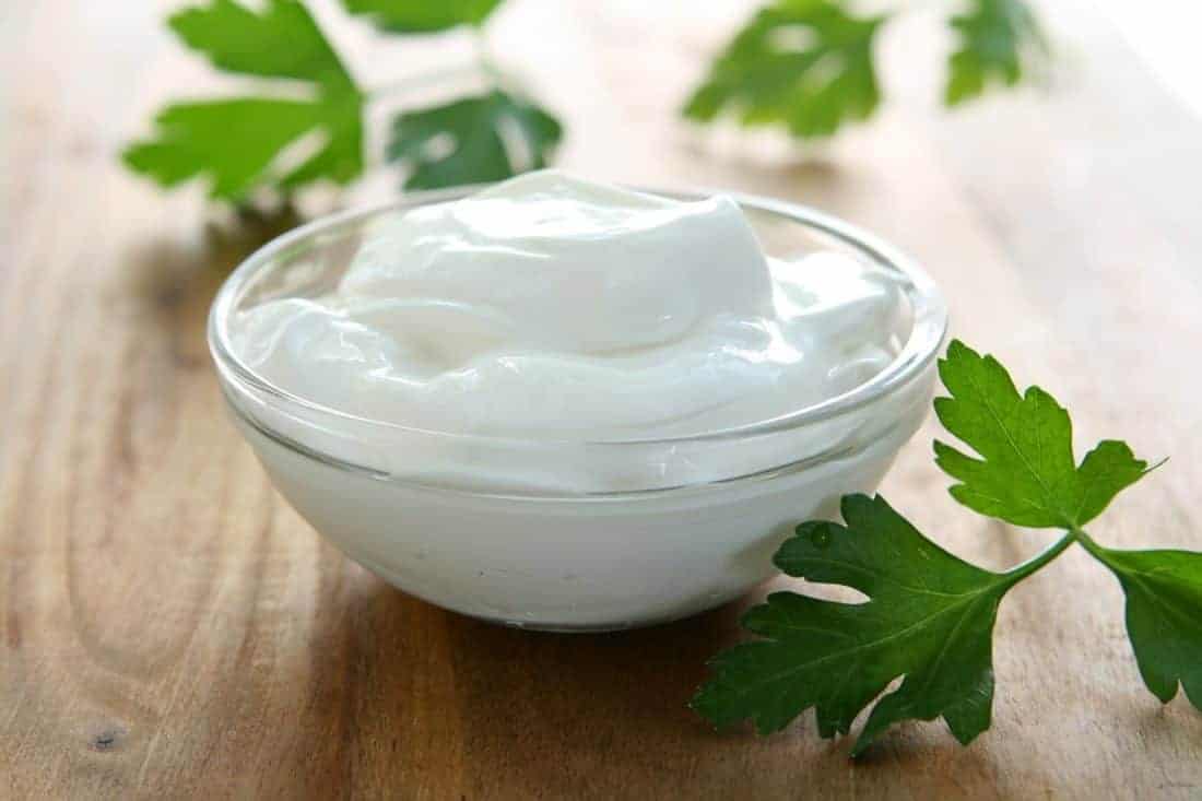 Sour Cream in a small transparent glass bowl on top of wooden table with some parsley leaves
