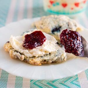 blueberry scones on a white plate cut in half and slathered with clotted cream and raspberry jam