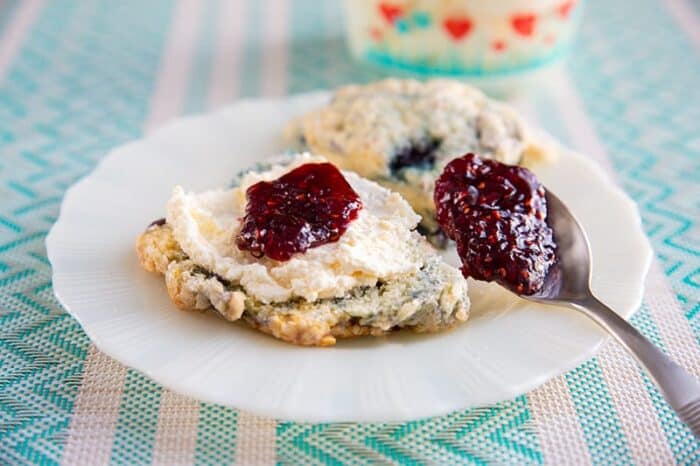 blueberry scones on a white plate cut in half and slathered with clotted cream and raspberry jam