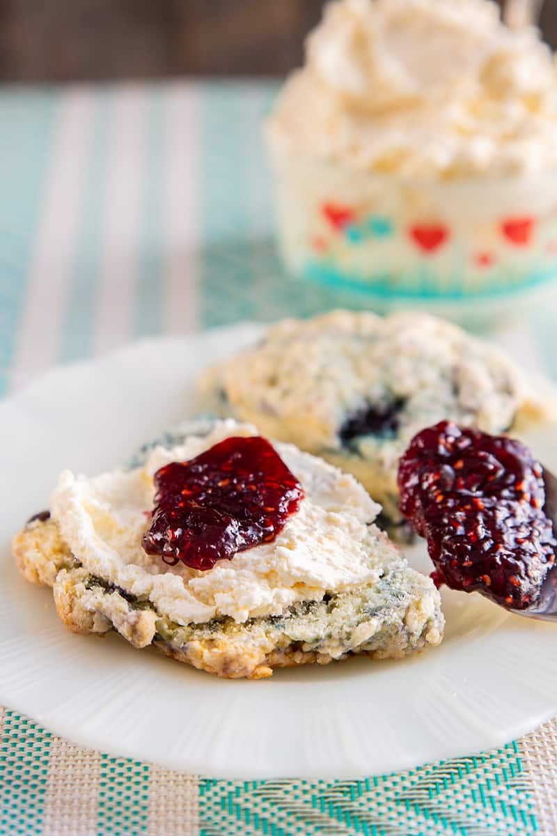 white plate with clotted cream on a scone topped with some jam, underneath is a pink and blue printed table cloth, clotted cream in a heart patterned Pyrex container in background