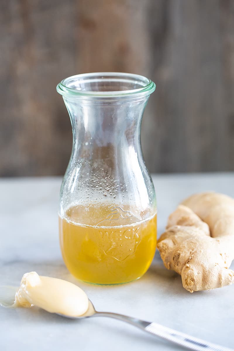 Ginger Simple Syrup in an airtight transparent glass with chuck of ginger beside it and a spoon of honey