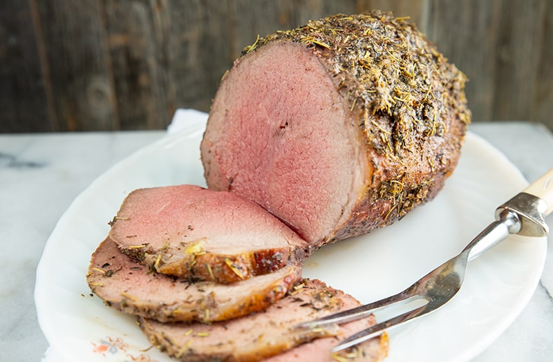 Eye of Round Roast with thin slices in a white plate with pot fork, stuffed with herbs and garlic