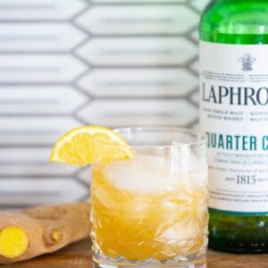 a glass of penicillin cocktail garnished with a lemon slice, a piece of ginger and a bottle of laphroaig