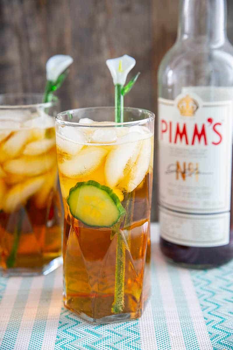 Pimm's cup cocktail in a tall glass with cucumber, and a glass lily flower stir stick.Bottle of Pimm's in background.