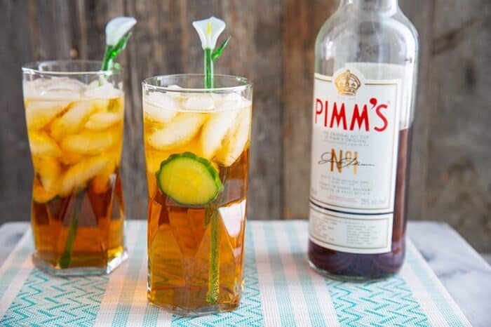 Two Pimm's cup cocktails in a tall glass with cucumber, and a glass lily flower stir stick.Bottle of Pimm's in background.