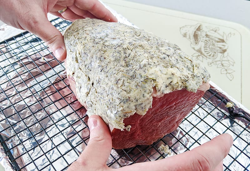 uncooked eye of round roast on a roasting rack slathered with butter herb mixture