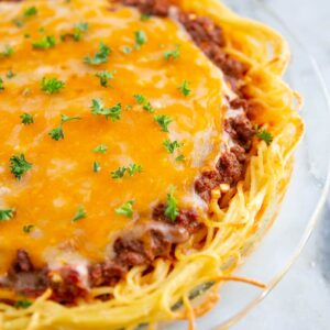 close up baked spaghetti pie on a large Pyrex plate with melted cheese on top and garnished with fresh parsley