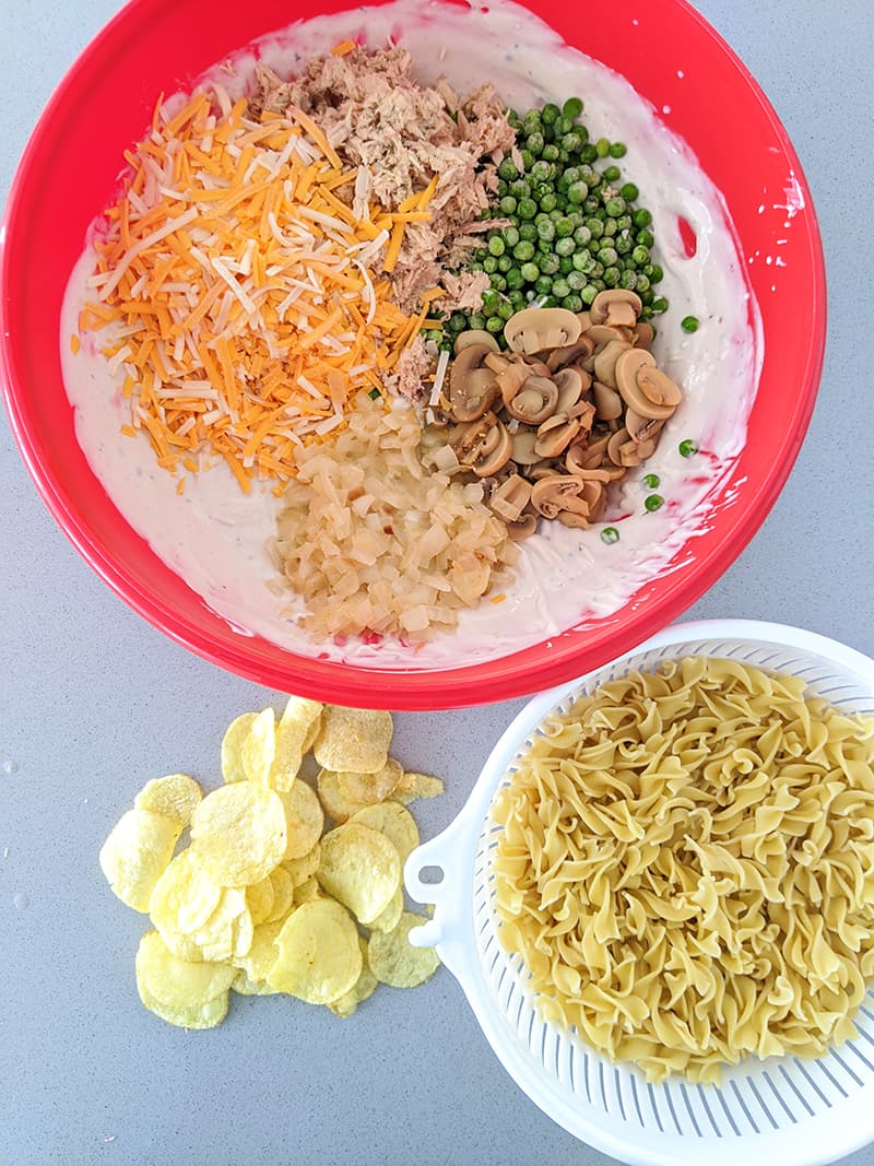 tuna casserole ingredients in a red Pyrex casserole egg noodles in a white strainer potato chips at the side