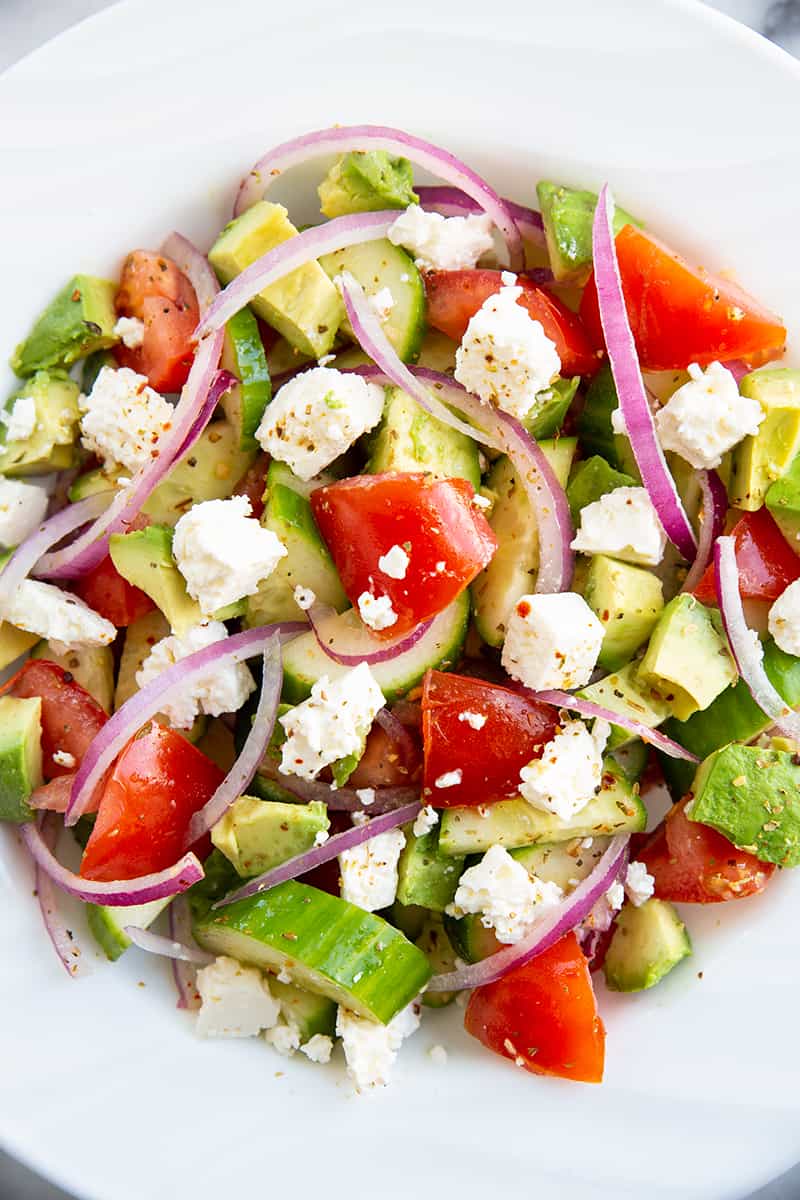 close up Greek avocado salad on a white bowl consists of sliced avocado, cucumber, tomatoes, lemon, feta, red onions and Greek seasoning