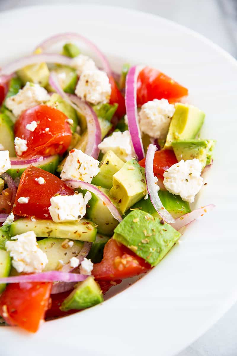 close up Greek avocado salad on a white bowl consists of sliced avocado, cucumber, tomatoes, lemon, feta, red onions and Greek seasoning