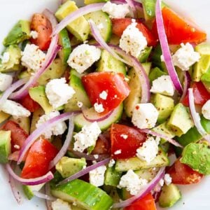 close up Greek avocado salad on a white bowl consists of sliced avocado, cucumber, tomatoes, lemon, feta, red onions and Greek seasoning