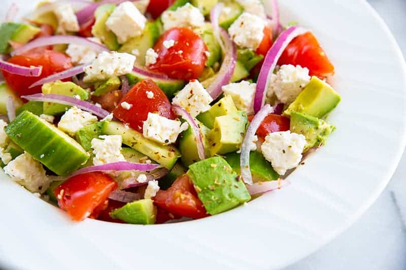 Greek avocado salad on a white bowl consists of sliced avocado, cucumber, tomatoes, lemon, feta, red onions and Greek seasoning