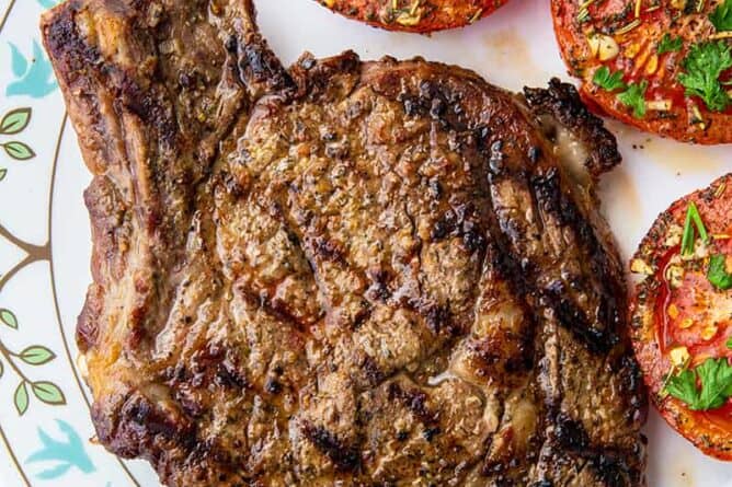 close up of grilled rib eye steak on a white plate with tomatoes