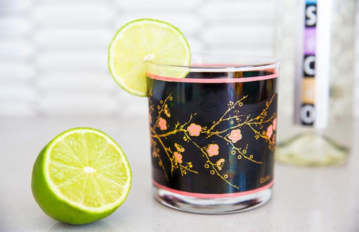 a lime beside a Pisco Sour Cocktail in a vintage glass that has pink and gold roses on a black background and a lime wedge,