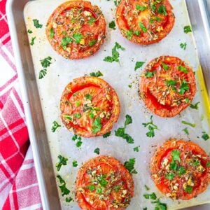 close up of oven roasted tomatoes on a sheet pan loaded with garlic and herbs on a red kitchen towel underneath