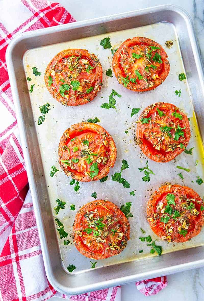close up of oven roasted tomatoes on a sheet pan loaded with garlic and herbs on a red kitchen towel underneath