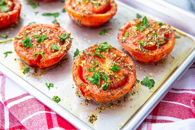 oven roasted tomatoes on a sheet pan loaded with garlic and herbs on a red kitchen towel underneath