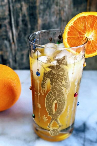 Vertical photo of a Brass Monkey Cocktail in a Mardis Gras style tumbler garnished with an orange. full orange in the background.