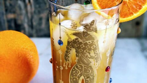 Vertical photo of a Brass Monkey Cocktail in a Mardis Gras style tumbler garnished with an orange. full orange in the background.
