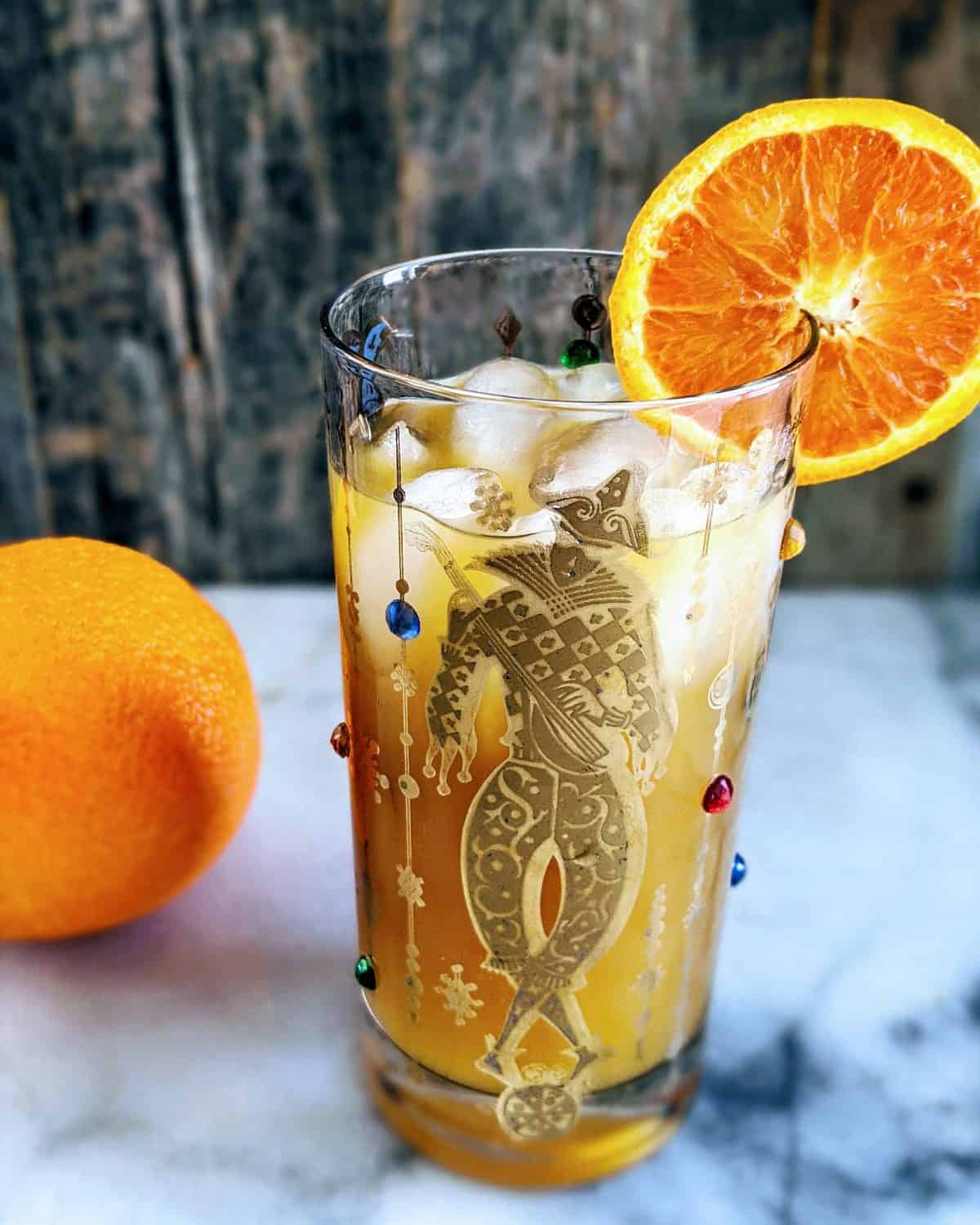 Vertical photo of a Brass Monkey Cocktail in a Mardis Gras style tumbler garnished with an orange. full orange in the background.