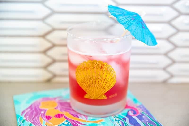 Bay Breeze cocktail in a glass emblazoned with a gold seashell garnished with a blue umbrella on a mermaid napkin.