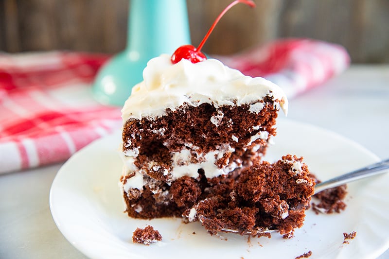 a slice of chocolate soda cake on a white plate with a piece of red cherry on top