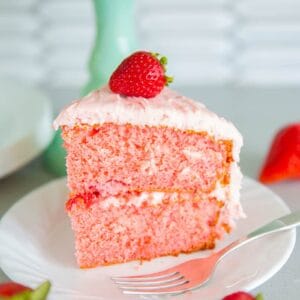 slice of strawberry cake on a white plate with a piece of fresh strawberry on top and a fork beside the plate