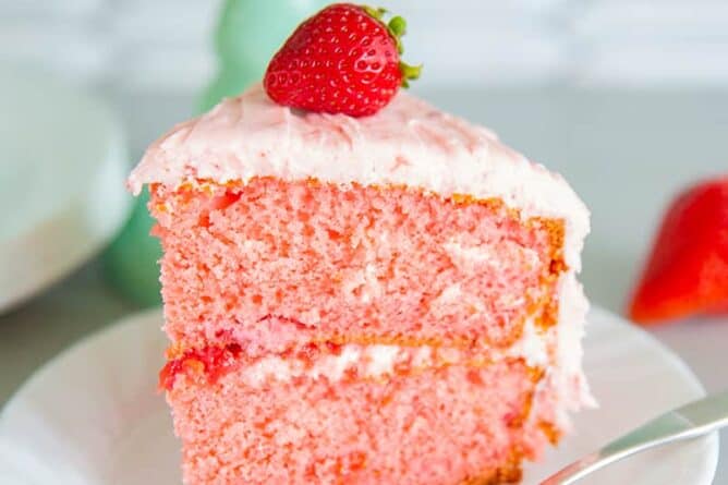 slice of strawberry cake on a white plate with a piece of fresh strawberry on top and a fork beside the plate