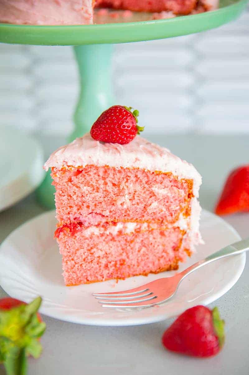 slice of strawberry cake on a white plate with a piece of fresh strawberry on top and a fork beside the plate
