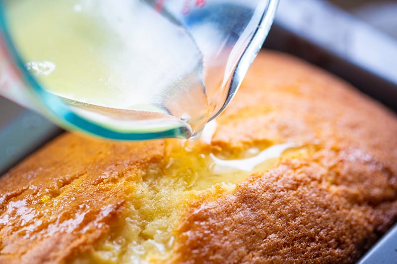 A close-up of a glass measuring cup pouring a clear glaze or syrup onto a golden-brown lemon bread loaf cake in a baking pan.