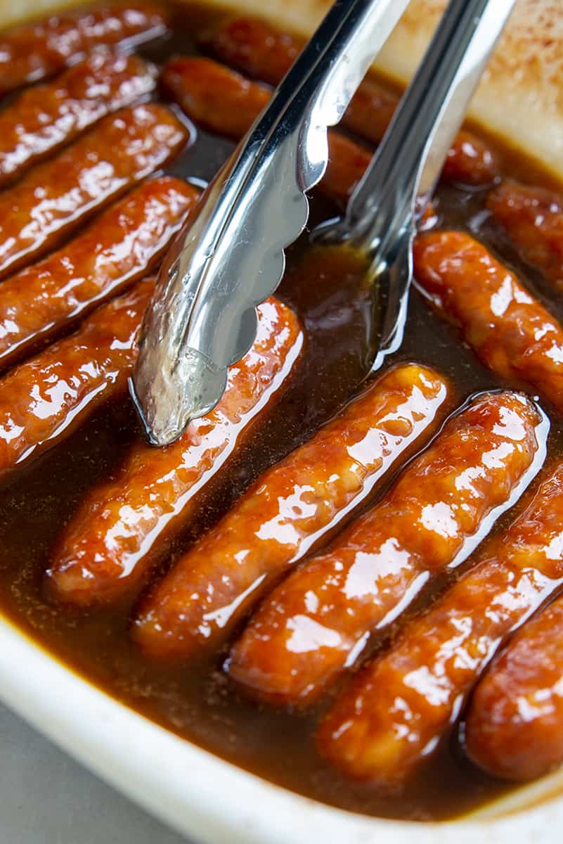 close up of pork breakfast sausages in a white dish in maple glaze syrup and a pair of metal tongs