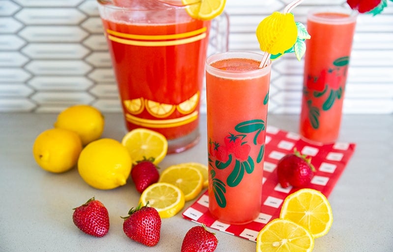 Strawberry Lemonade in two glasses and a pitcher with lemons and strawberries on a counter