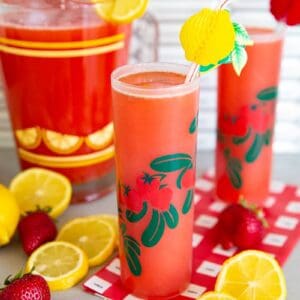 Strawberry Lemonade in two glasses and a pitcher with lemons and strawberries on a counter