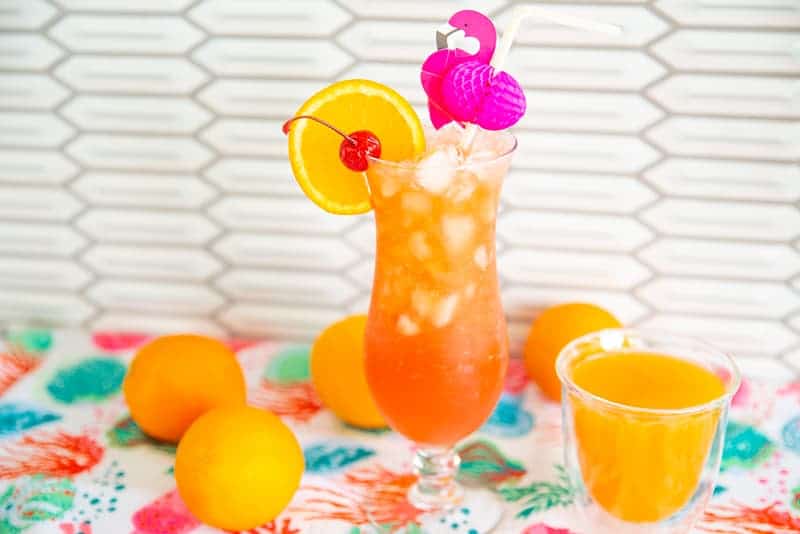 Hurricane Cocktail in a hurricane glass on a counter top surrounded by oranges with a side of passion fruit juice.