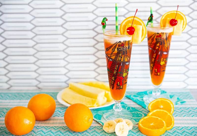 Rum Runner Cocktail in a tall pirate designed pilsner glass on a turquoise tablecloth surrounded by oranges, pineapple wedges and banana slices.