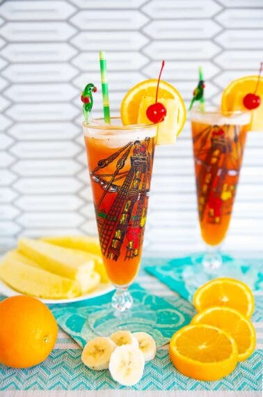 Rum Runner Cocktail in a tall pirate designed pilsner glass on a turquoise tablecloth surrounded by oranges, pineapple wedges and banana slices.